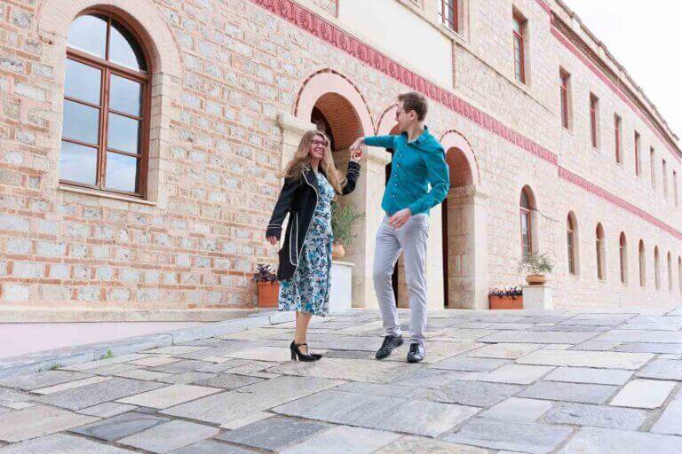 A happy couple dancing and laughing on a cobblestone street in Athens, with a beautiful historic stone building in the background during a professional photo session.